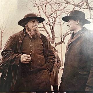 Two men in th century clothing outdoors wearing wide brimmed hats one with a beard holds a pipe the other in a dark coat
