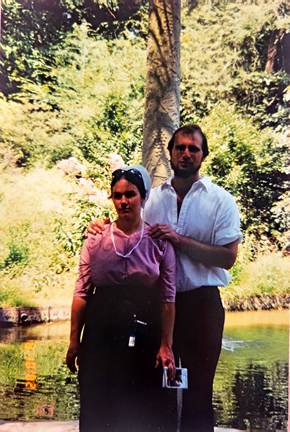 Couple standing together in a sunny park by a pond with trees behind them posing for a photo