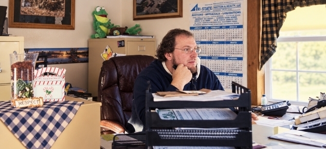 Man in a navy hoodie sits at a cluttered office desk resting his chin on his hand while looking to the right